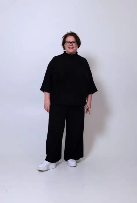 Der Hintergrund ist grau. in der Mitte steht eine Frau mit kurzen rötlichem Haar. Sie ist ganz in Schwarz gekleidet.