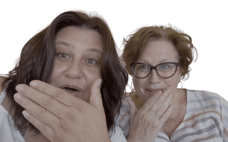 Zwei Frauen mit überraschter Mimik schauen direkt in die Kamera, beide mit der Hand vor dem Mund.