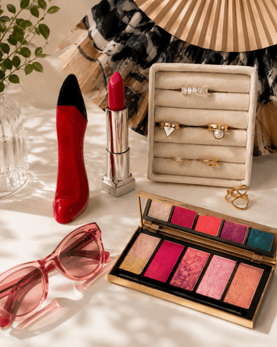 Goldschmuck mit Ringen, Ketten und Armband auf einer hellen Schmuckablage, kombiniert mit rotem Lippenstift, pinker Sonnenbrille und farbiger Make-up-Palette.